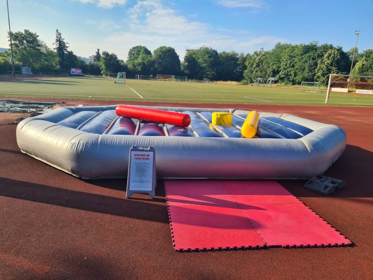Ein aufblasbares Sportgerät auf einem Roten Teppich vor einem Sportfeld.