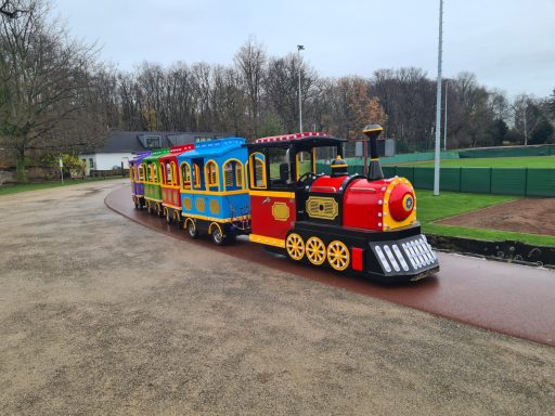Bunte Miniatur-Dampflok mit mehreren Waggons auf einem Radweg.