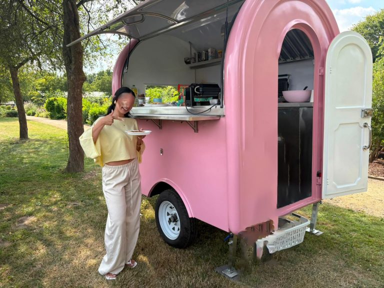 Frau steht neben einem pinken Food-Truck in einer grünen Parklandschaft.