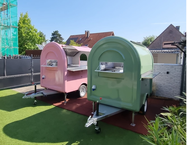 Zwei farbige Food-Trucks, pink und grün, stehen auf einem Rasen vor einem Gebäude.