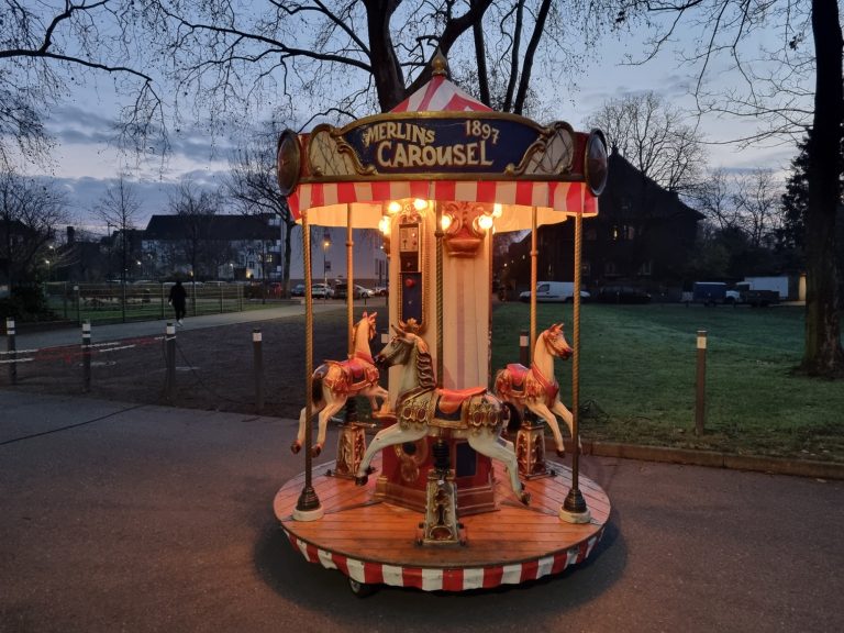 Merlins Karussell Karussell mit beleuchteten Pferden in einem parkähnlichen Bereich bei Dämmerung.