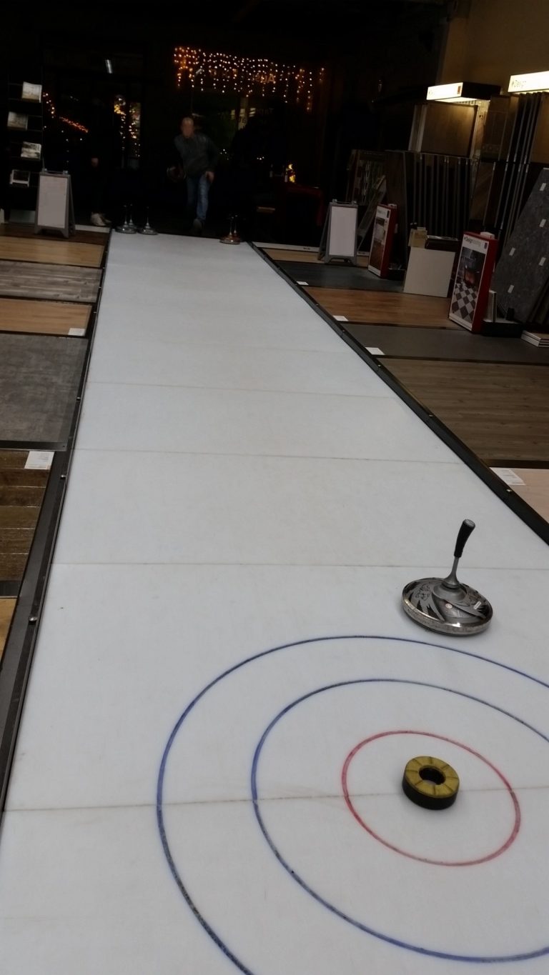 Eine Curlingbahn mit einem Stein auf dem Zielring und Zuschauern im Hintergrund.