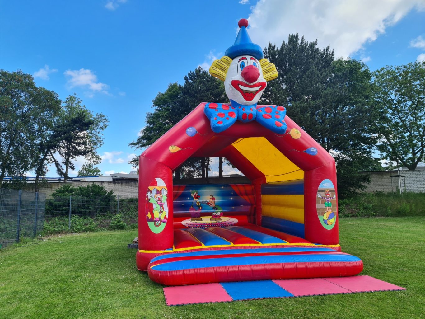 Hüpfburg Clown - Verschiedene Ausführungen Bunte Hüpfburg mit Clownsgesicht, auf einer grünen Wiese unter blauem Himmel.