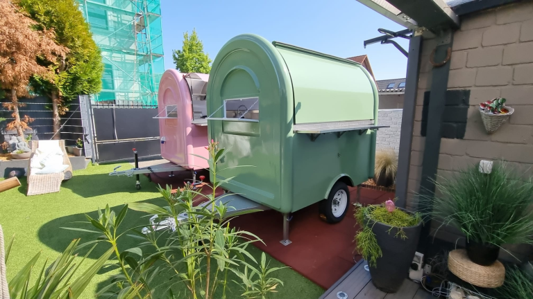 Ein grüner Wohnwagen steht in einem Garten mit Pflanzen und einem pinken Anhänger.